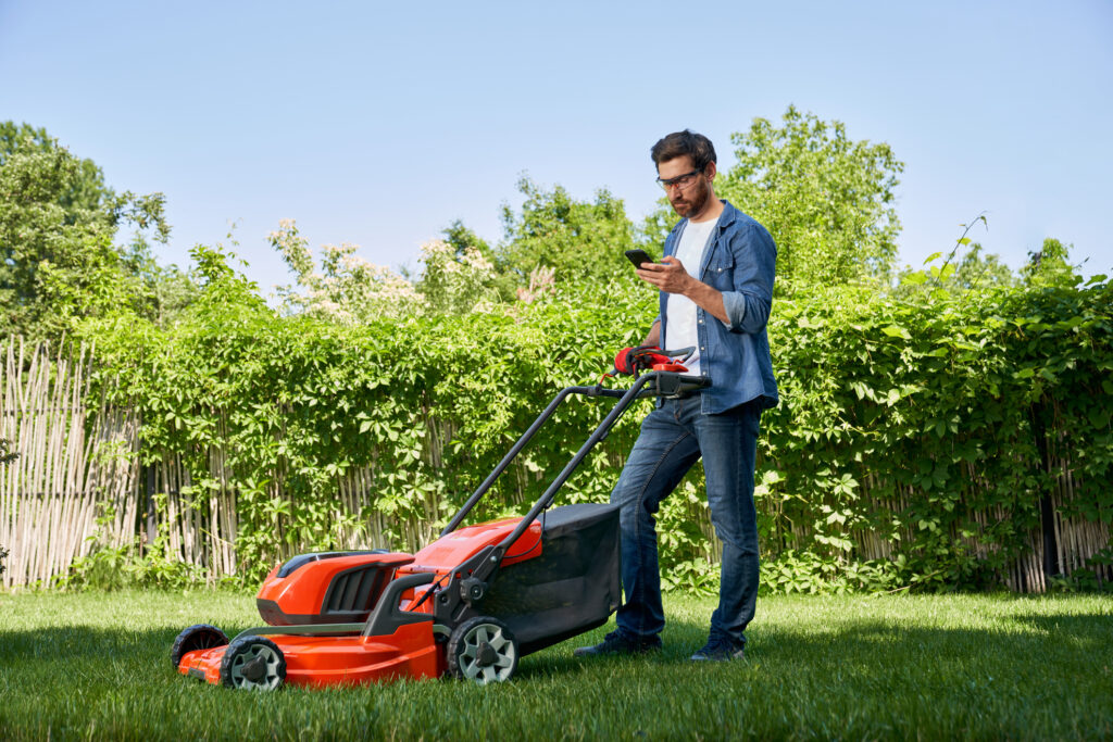 Focused male gardener using smartphone, while having break after trimming lawn in garden.