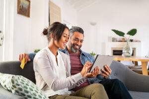 Happy multiethnic mature couple using digital tablet for online payment with credit card. Cheerful latin wife showing something to buy on digital tablet to her indian husband while holding bank card. Mid adult hispanic woman and middle eastern man sitting on couch in living room while doing shopping online together.