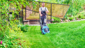 The gardener takes care of the lawn by mowing the grass with an electric lawn mower. A man surfs the Internet and engages in online trading during chores and gardening. Gardener services concept.